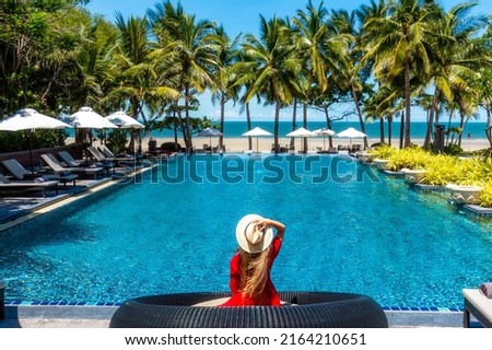 Luxury beach vacation in tropical beach hotel. Tourist woman in red dress relax near blue swimming pool in modern resort. Female traveler on sea vacation. Royalty-Free Stock Photo #2164210651