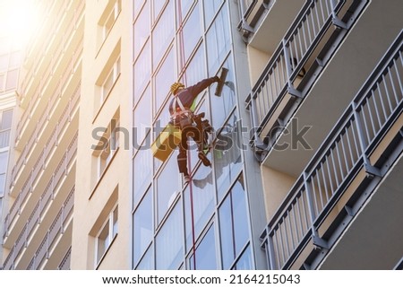 Concept of industry urban works. Industrial mountaineering worker in uniform hangs over residential facade building, washing exterior glazing. Rope access laborer hangs on wall of house. Copy space Royalty-Free Stock Photo #2164215043