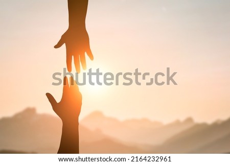 Silhouette of two people hands reaching to each other for help in sunset sky and orange sun. Friendship, teamwork, help, faith and hope concept. Royalty-Free Stock Photo #2164232961