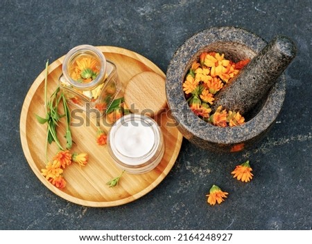 Calendula Cream for body care in glass jar and fresh orange calendula flowers on a wooden plate and a granite mortar on black table. Concept: spa, aromatherapy, Medical Dermatology. Flat lay