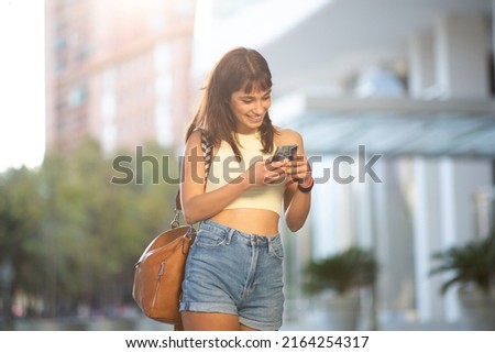 Portrait of smiling young woman walking outside in the city looking at mobile phone