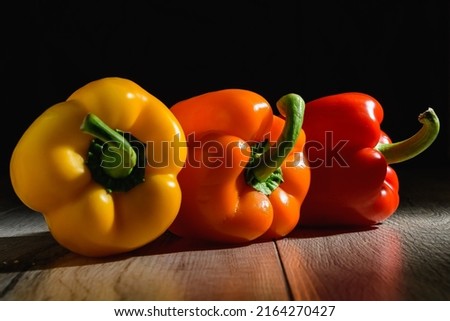 The three colorful bell peppers are on the wooden table. There is dark photography of yellow, red, and orange capsicums.