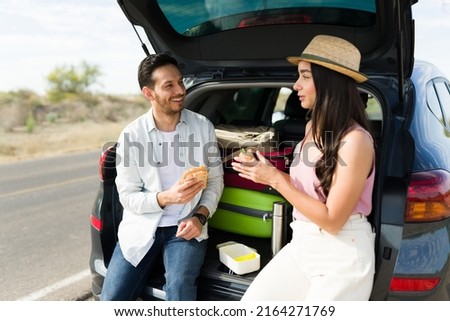 Attractive young man and woman eating a sandwich while resting on the car trunk during a weekend trip together Royalty-Free Stock Photo #2164271769