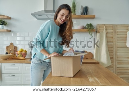 Beautiful young woman unpacking box while standing at the domestic kitchen Royalty-Free Stock Photo #2164273057