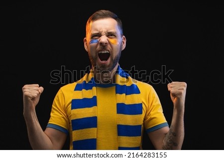 Portrait of euphoric happy soccer supporter man in yellow-blue t-shirt and scarf and painted face, cheering for his favourite team, isolated over black background. Royalty-Free Stock Photo #2164283155