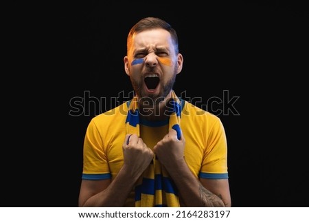 Portrait of excited young soccer supporter man in yellow-blue t-shirt and scarf and painted face, cheering for his favourite team. Isolated over black background. Royalty-Free Stock Photo #2164283197