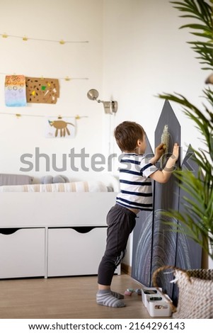 Cute baby boy drawing on black chalkboard use colorful chalks indoor at childish playroom kindergarten. Funny playful male kid creative art picture use crayon at home. Adorable baby happy childhood