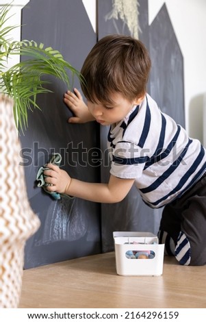 Adorable male kid wiping black chalkboard with crayon creative drawing picture use rag tidying up cleaning playroom. Cute baby boy maintaining clean at home doing housework at kindergarten room