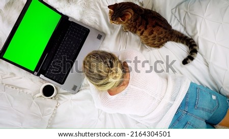 Top-down view of a woman lying on the bed looking at a blank green screen of a laptop computer drinking coffee, Bengal cat sitting nearby