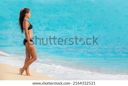 Asian bikini body woman going swimming in Caribbean ocean on beach vacation. Beautiful swimsuit model with lean legs and body profile wearing black swimwear on blue background Royalty-Free Stock Photo #2164332521