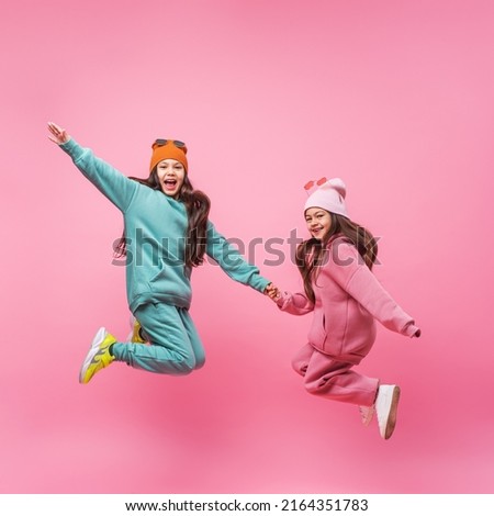 Two smiling Latin child girls in warm fashionable tracksuits jumping on a pink studio copy space. Children's beauty models with dark long hair advertising sports outerwear. Promotion of clothing store