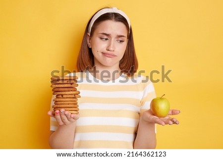 Indoor shot of puzzled confused woman wearing t shirt and hair band posing isolated on yellow background, holding cookies and apple, doesn't know what to choose, healthy or junk food.