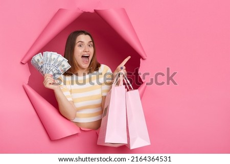 Portrait of excited surprised attractive woman wearing striped shirt looking through breakthrough of pink background, pointing finger aside, holding fan of dollars and shopping bags.