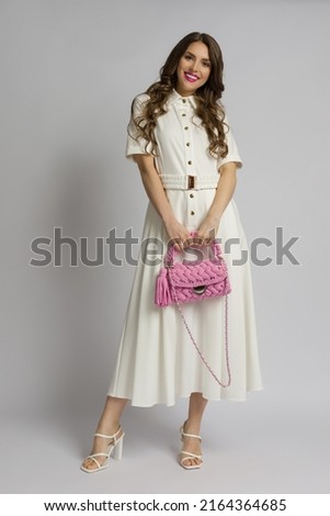 Young woman in white maxi dress and high heels is  holding a pink braided purse. Front view. Full length studio shot. Royalty-Free Stock Photo #2164364685