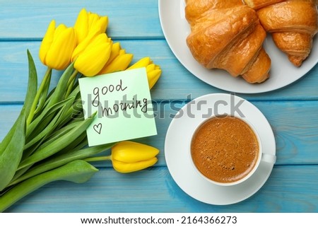 Cup of aromatic coffee with croissants, beautiful yellow tulips and Good Morning note on light blue wooden table, flat lay