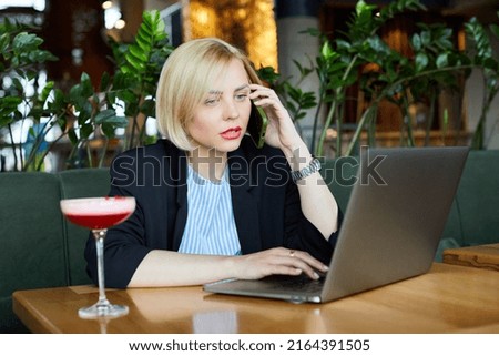 Picture of inspired attractive model writing at her laptop, spending time alone in cafe, having glass of cocktail on table, being busy, wearing suit. People and activities concept.