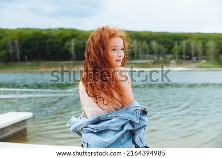 a little beautiful girl with red long hair stands on the shore of the lake. romantic portrait