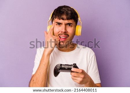 Young hispanic man playing with a video game controller isolated on purple background showing a disappointment gesture with forefinger.