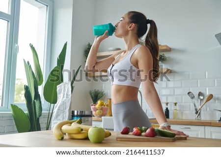 Beautiful young woman in sports clothing drinking protein cocktail while standing at the kitchen Royalty-Free Stock Photo #2164411537