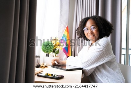 Working woman LGBTQ+ Afro hair style African American lesbian, Beautiful gay working on tablet with love moment spending good time together, lgbt rainbow, pride flag on table near curtain at window. Royalty-Free Stock Photo #2164417747
