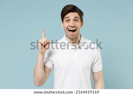Young smart proactive excited happy unshaven caucasian man 20s wearing white casual basic t-shirt holding index finger up with great new idea isolated on pastel blue color background studio portrait.