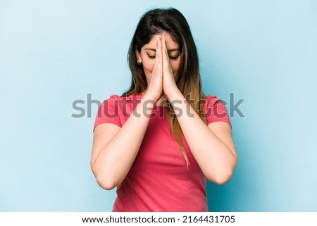 Young caucasian woman isolated on blue background holding hands in pray near mouth, feels confident.