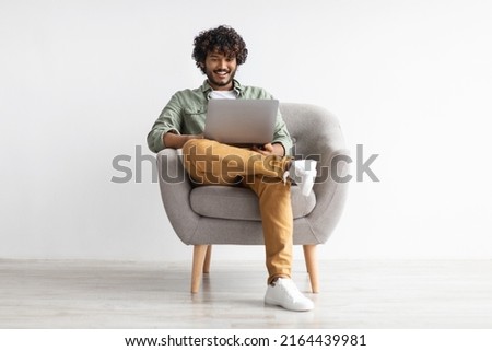 Joyful young indian guy in casual sitting in armchair, chilling with modern laptop, gambling or gaming online, surfing on Internet, white studio background, full length shot, copy space Royalty-Free Stock Photo #2164439981