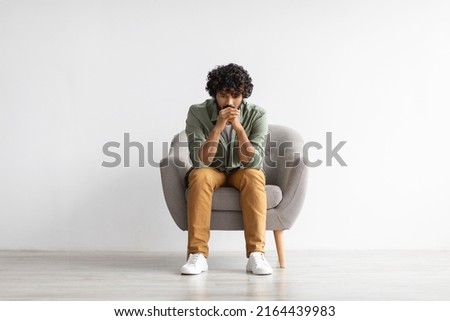 Depressed curly bearded young indian guy in casual outfit sitting in armchair over white studio background, looking down, thinking about something, suffering from loneliness, copy space, full length Royalty-Free Stock Photo #2164439983