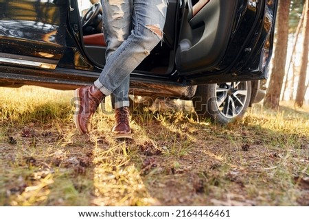 View of jeans and shoes. Man in jeans is outdoors in the forest with his black colored automobile.