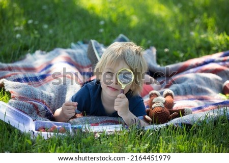Cute toddler child, blond boy, playing with magnifying glass  and drawing in a picture book in the park, summertime