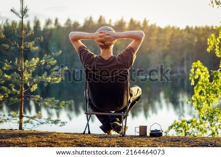 A man is sitting in a camping chair on a summer evening on the background of a forest lake. Object in focus, background blurred.