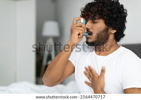 Indian guy suffering from asthma, using inhaler in bed, side view, copy space. Young hindu man woke up with asthma attack, touching chest and using nebulizer, bedroom interior Royalty-Free Stock Photo #2164470717