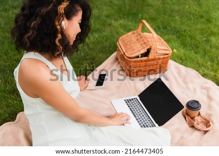 curly african american woman using laptop near wicker basket and smartphone on blanket in park