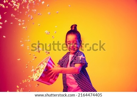 Flying popcorn. One happy little girl, pupil wearing 3d glasses and holding bucket of popcorn isolated on orange background in neon. Concept of emotions, facial expression, youth, aspiration
