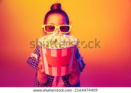 Closeup. One happy little girl, pupil wearing 3d glasses and holding bucket of popcorn isolated on orange background in neon. Concept of emotions, facial expression, youth, aspiration