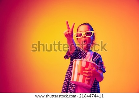 Victory sign. One happy little girl, pupil wearing 3d glasses and holding bucket of popcorn isolated on orange background in neon. Concept of emotions, facial expression, youth, aspiration