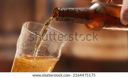 Beer is poured from dark brown bottle into beer glass. Close-up light fresh beer poured into glass steamed up from cold. Lager beer foams and pours from bottle into glass. Royalty-Free Stock Photo #2164475175