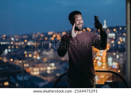 Cheerful young man taking selfie on balcony with urban view at night.