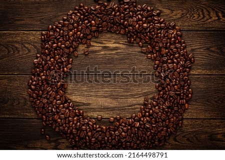 Roasted coffee beans arranged into ring. Brown grains scattered in circle over dark wooden board background with empty space in middle. Round frame top view