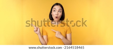 Portrait of intrigued asian woman, looking and pointing fingers left at advertisement, showing smth interesting, standing over yellow background