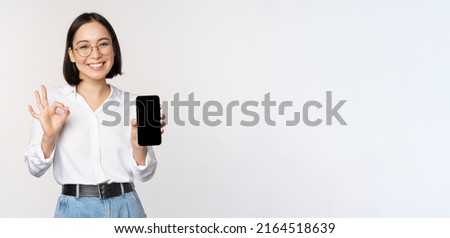 Image of asian businesswoman, showing smartphone screen, app interface and ok sign, recommending application on mobile phone, standing over white background