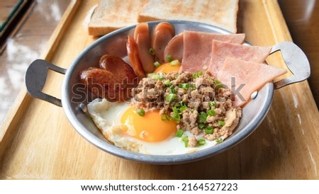 Fried eggs in a small pan with ham, minced pork, spring onion and chinese sausages on wood tray. Popular Thai Breakfast called Kai Kata.