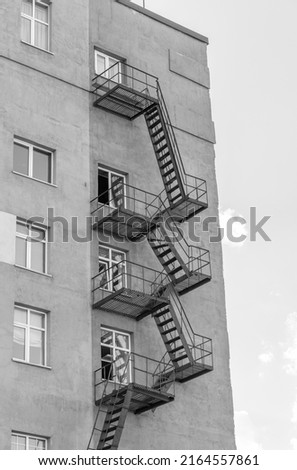 Silhouette of a fire escape on a high-rise building against a blue sky with clouds. Some of the stairs are broken. There is free space for text. Black and white photo