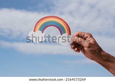 A paper-cut cute rainbow in a man's hands on a beautiful blue sky and white cloud in sunny day with natural light, Positive picture or happy  life or World mental health day and eco friendly concept.