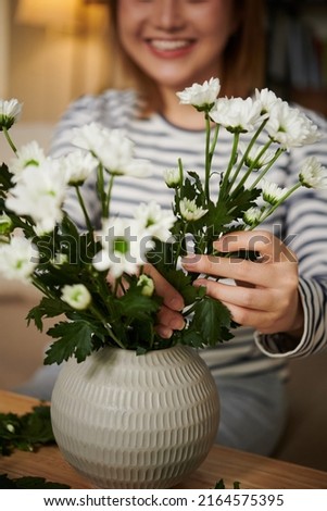 Close-up image of smiling young woman putting chrysanthemum flowers in short vase