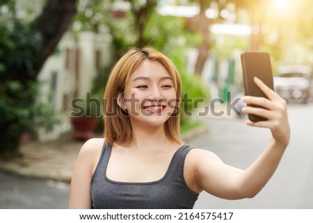 Pretty smiling young Asian woman taking selfie outdoors after morning run