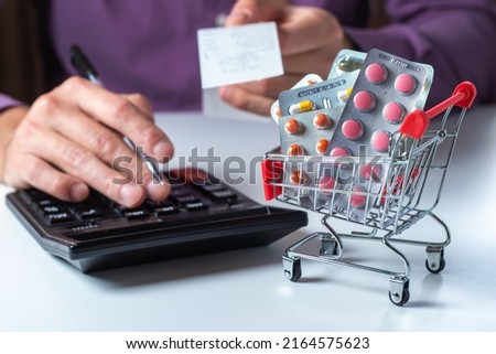 Concept of large spending on medicines. Shopping cart full of medicines and pills. A man calculates the cost of medicines on a calculator holding a check in his hand. Royalty-Free Stock Photo #2164575623