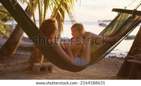 Young mother sitting with her daughter in a hammock on the beach laughing and smiling against the background of the sunset. Happy family relaxing in tropical hammock at sunset. Summer luxury vacation