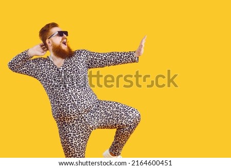 Funny eccentric overweight man dressed in leopard print pajamas dancing and fooling around on orange background. Cheerful bearded fat man in sunglasses having fun at copy space. Web banner.