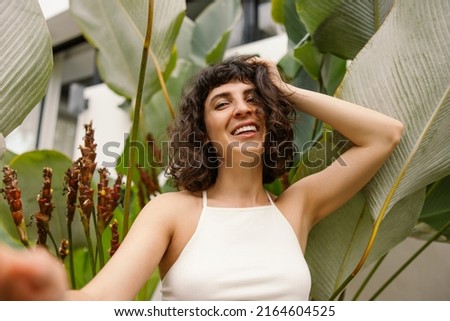 Attractive young caucasian girl posing smiling at camera against backdrop of tropical plants. Brunette with short haircut wears white top. Summer vacation concept